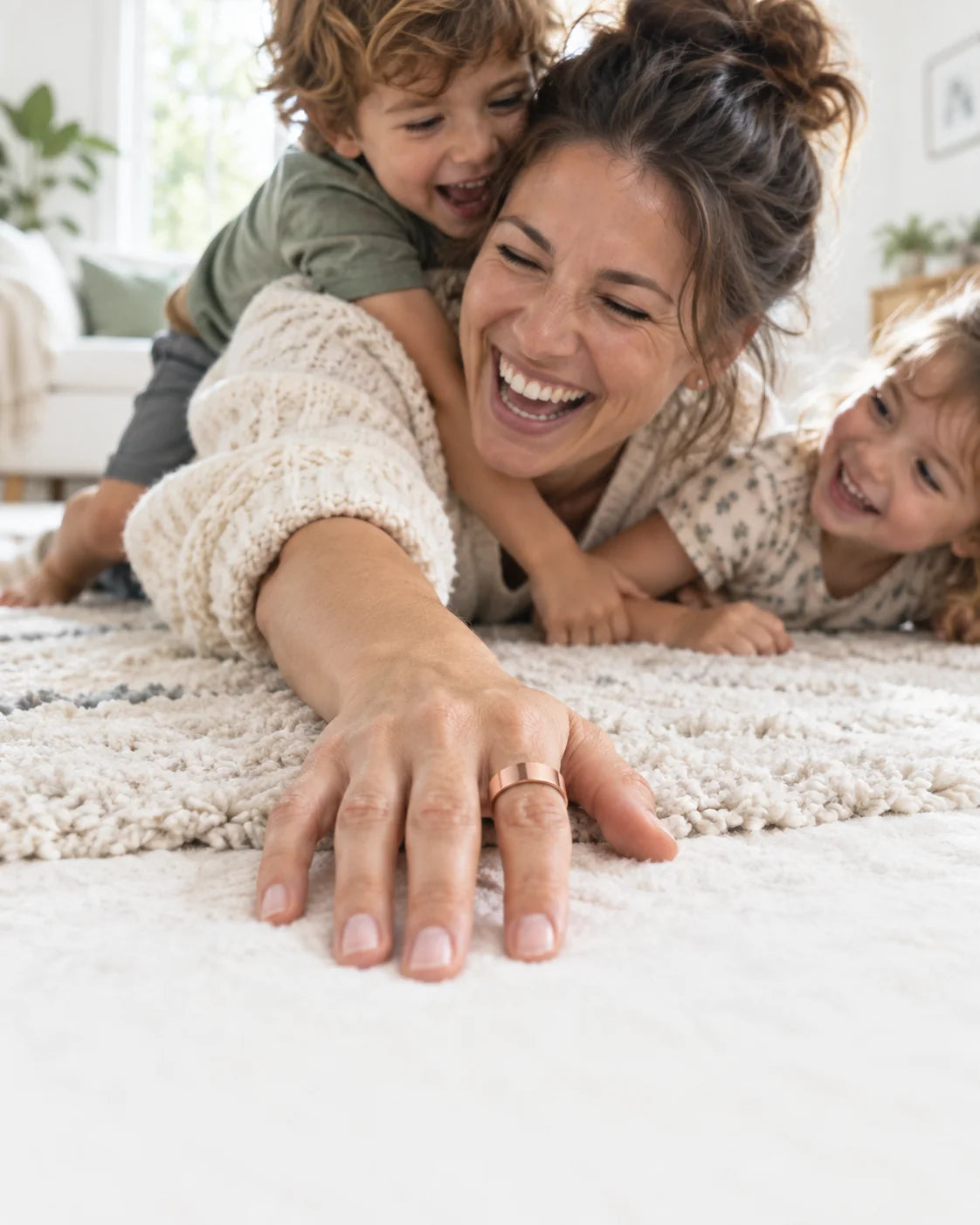 A mother laughs on the living-room rug with her two kids, the Vitality Ring on her hand