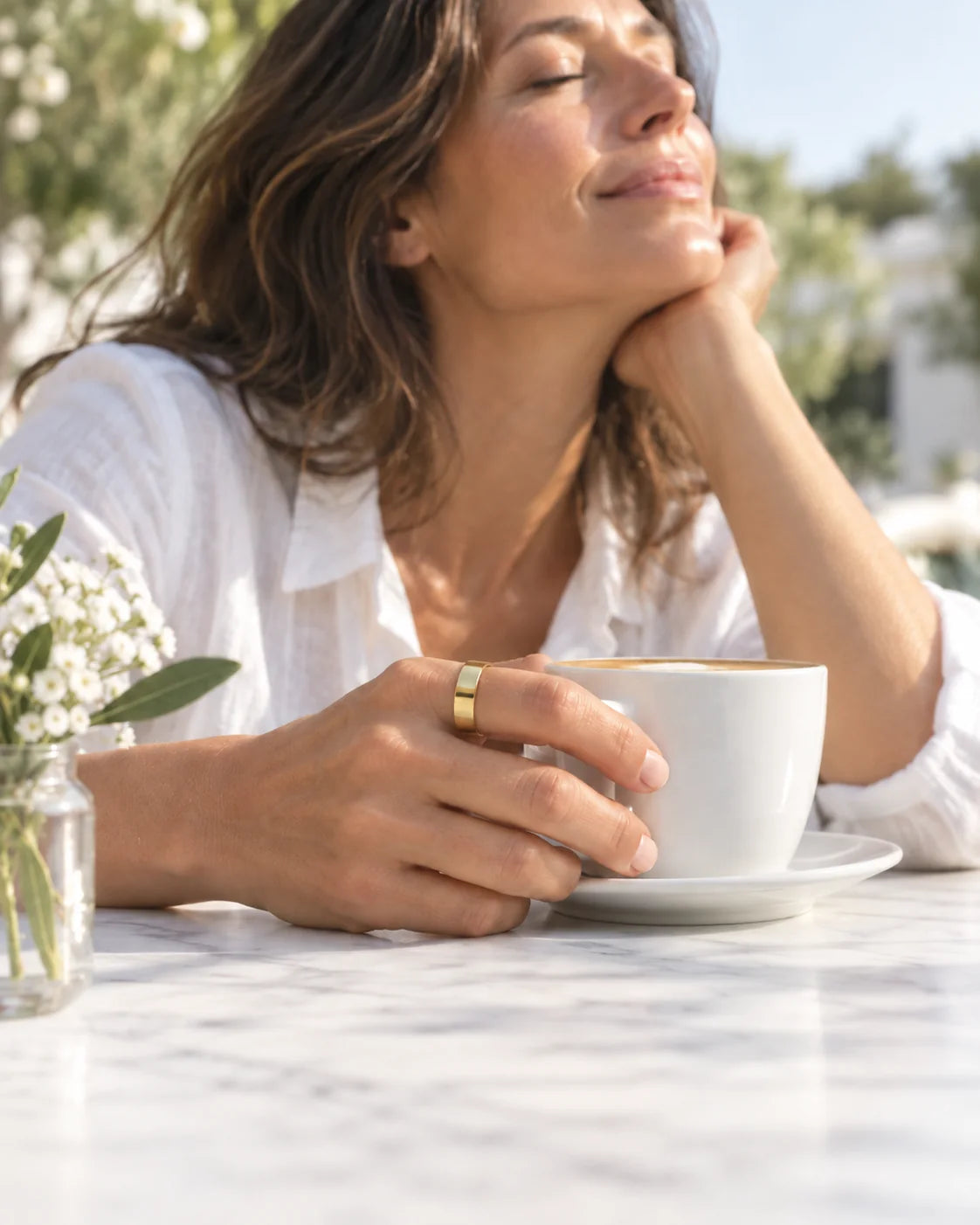 A woman relaxes on a sunny café terrace with a coffee, eyes closed, the Vitality Ring on her hand