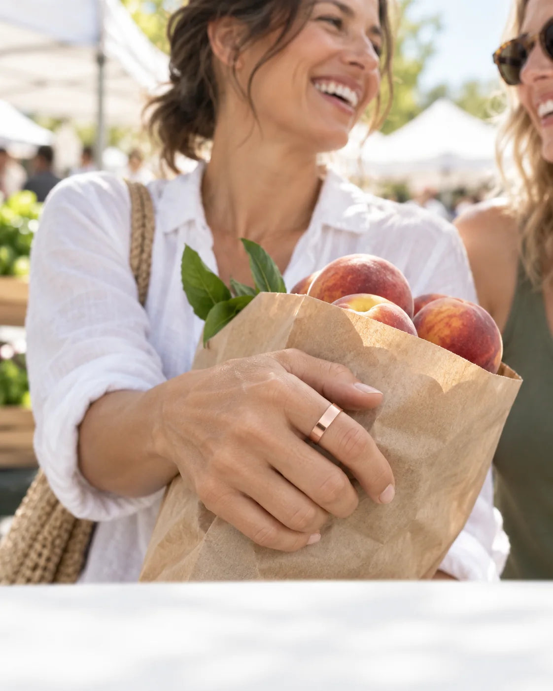 A woman laughs with a friend at a Saturday market, holding a bag of peaches, the Vitality Ring on her hand