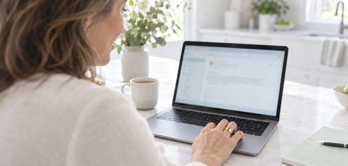 Over the shoulder of a woman reading an email on her laptop at a white kitchen table, the Vitality Ring on her hand